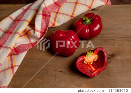 Two whole and halves of red bell pepper on wooden background with red kitchen towel. . Two whole and halves of red bell pepper on wooden background with red kitchen towel. . 114365050