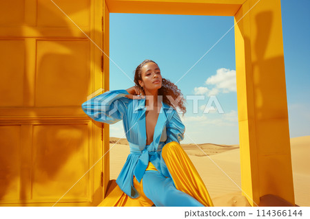 A woman in a blue jacket and yellow pants sits in front of a yellow door 114366144