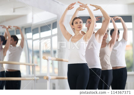 Beautiful graceful women and sports men practice at ballet standing in fifth position at barre in choreography studio Beautiful graceful women and sports men practice at ballet standing in fifth position at barre in choreography studio 114367371