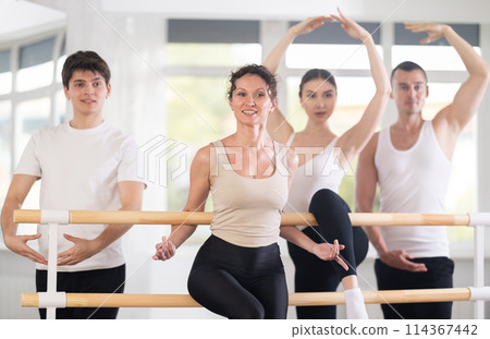 Beautiful young woman ballerina posing with group of dancers in ballet training hall Beautiful young woman ballerina posing with group of dancers in ballet training hall 114367442