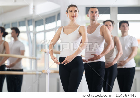 Positive girl with women and men engaged in group class perform exercise near ballet barre, standing in first position of ballet stance in dance studio Positive girl with women and men engaged in group class perform exercise near ballet barre, standing in first position of ballet stance in dance studio 114367443