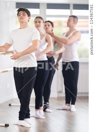 Group of women and men standing in row together while doing ballet dance moves near ballet barre in dancing hall, male choreographer helps to coordinate movements correctly 114367581