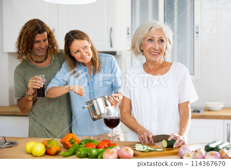 Happy couple drinking wine while mature woman cooking vegetable salad at cosy kitchen 114367670