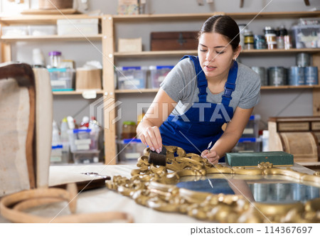 Woman restorer restores wooden frame surface of vintage mirror in workshop 114367697