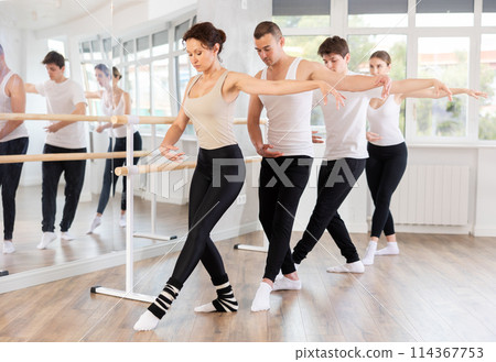 Group of positive women and men standing in row together while doing ballet dance moves dancers standing with one hand on barre and reaching over hand in fitness room Group of positive women and men standing in row together while doing ballet dance moves dancers standing with one hand on barre and reaching over hand in fitness room 114367753