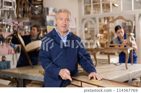 Elderly experienced restorer man carefully and carefully restores chest of drawers in restoration workshop 114367769