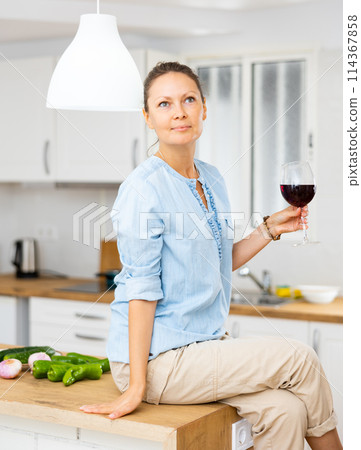 Positive casual lady relaxing with red wine and sitting with crossed legs at modern kitchen 114367858