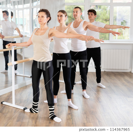 Dancing positive women and men engaged in group class perform exercise near ballet barre, standing in 2nd position of ballet stance in dance studio Dancing positive women and men engaged in group class perform exercise near ballet barre, standing in 2nd position of ballet stance in dance studio 114367965