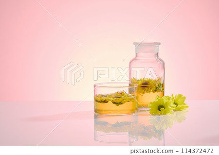 A glass bottle with yellow liquid and calendula in, placed next to a glass cup, decorated by fresh flowers on the right side, over light pink background. Blank left side for displaying, front view A glass bottle with yellow liquid and calendula in, placed next to a glass cup, decorated by fresh flowers on the right side, over light pink background. Blank left side for displaying, front view 114372472
