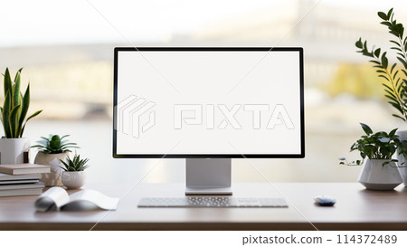 A computer white-screen mockup, a keyboard, books, and potted plants on a desk against the window. A computer white-screen mockup, a keyboard, books, and potted plants on a desk against the window. 114372489