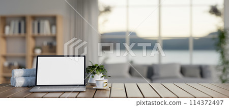 A wooden table features a white-screen laptop computer mockup in a contemporary living room. A wooden table features a white-screen laptop computer mockup in a contemporary living room. 114372497
