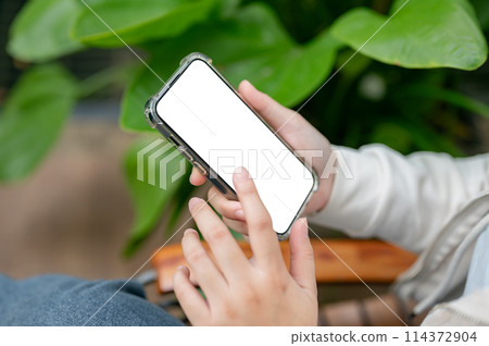A close-up image of a woman is sitting next to a houseplant indoors and using her smartphone. 114372904