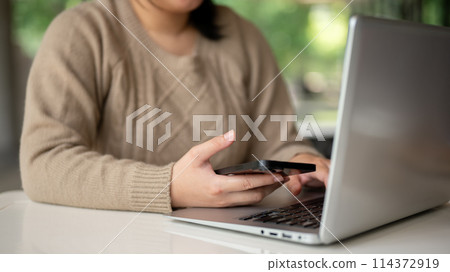 A woman holding a smartphone while typing on her laptop keyboard, working on her computer at a table 114372919