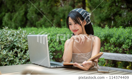 An attractive woman is working remotely in a garden, looking at her laptop, satisfied with her work. 114374409
