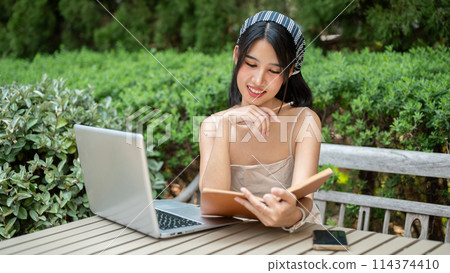 A beautiful Asian woman is working remotely or studying online in a garden, reading a book. 114374410