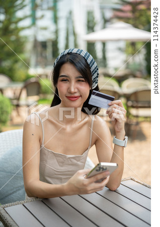 An attractive Asian woman sits at a table in a garden holding a credit card and smartphone. An attractive Asian woman sits at a table in a garden holding a credit card and smartphone. 114374428