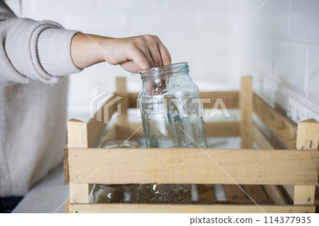 A woman wants to send washed glass jars for recycling and puts them in a drawer in the kitchen. An eco activist sorts garbage at home, taking care of environmental pollution. Garbage recycling. A woman wants to send washed glass jars for recycling and puts them in a drawer in the kitchen. An eco activist sorts garbage at home, taking care of environmental pollution. Garbage recycling. 114377935