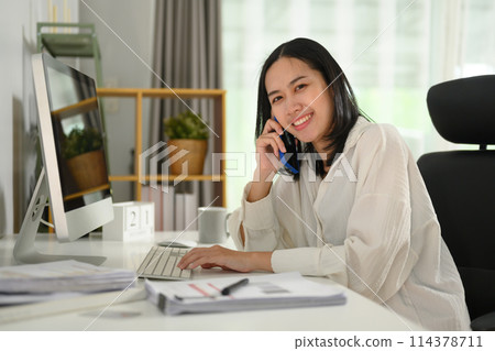 Beautiful businesswoman sitting at working desk and talking on mobile phone 114378711