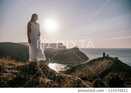 A woman stands on a rocky hill overlooking the ocean. She is wearing a white dress and she is enjoying the view. The scene is serene and peaceful, with the sun shining brightly in the background. A woman stands on a rocky hill overlooking the ocean. She is wearing a white dress and she is enjoying the view. The scene is serene and peaceful, with the sun shining brightly in the background. 114378821