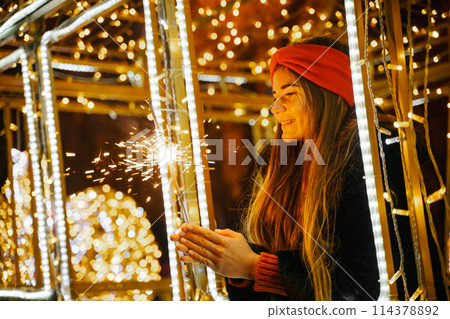 Woman holding sparkler night while celebrating Christmas outside. Dressed in a fur coat and a red headband. Blurred christmas decorations in the background. Selective focus Woman holding sparkler night while celebrating Christmas outside. Dressed in a fur coat and a red headband. Blurred christmas decorations in the background. Selective focus 114378892