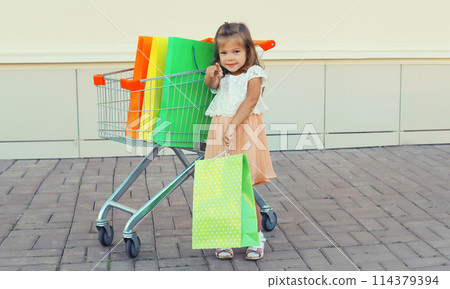 Happy little girl child with trolley cart and colorful shopping bags on city street 114379394