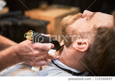 Close-up of professional barber trimming client's beard. Male hairdresser serving client with thin beard by clipper. Isolated photo of man getting his haircut done. 114380697