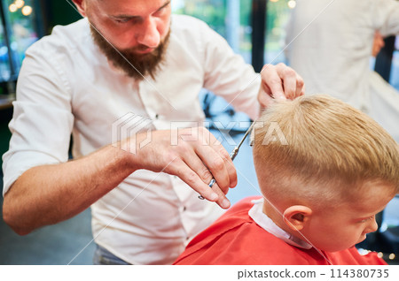 Man barber cutting little boy's hair using comb and scissors. Child getting haircut from adult male, likely barber. Professional hairdresser and cute client at modern barbershop. Man barber cutting little boy's hair using comb and scissors. Child getting haircut from adult male, likely barber. Professional hairdresser and cute client at modern barbershop. 114380735