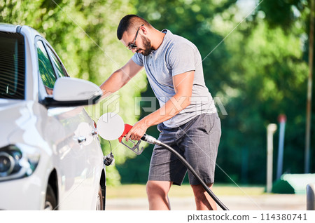 Driver with gasoline pump refilling car gas tank. Confident man refueling his luxury white auto. Man in casual clothes and sunglasses at modern gas station. 114380741