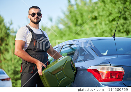 Stalled car filling with gasoline by professional worker. Portrait of young man refueling gas tank with fuel from canister. Male worker in overalls and sunglasses filling blue car. Stalled car filling with gasoline by professional worker. Portrait of young man refueling gas tank with fuel from canister. Male worker in overalls and sunglasses filling blue car. 114380769