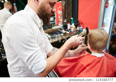 Barber using comb and shaver to cut hair. Professional hairdresser shaving little kid's nape. 114380777