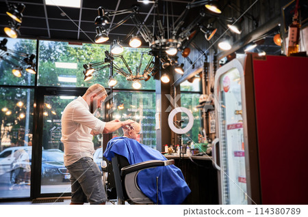 Man barber cutting little boy's hair using comb and scissors. Child getting haircut from adult male, likely barber. Professional hairdresser and cute client at modern barbershop. Man barber cutting little boy's hair using comb and scissors. Child getting haircut from adult male, likely barber. Professional hairdresser and cute client at modern barbershop. 114380789