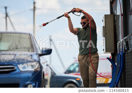 Confident man washing his car on self car-washing station. Cleaning car using active foam. Young man cleaning his auto by water gun. 114380815