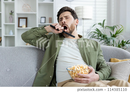 A bored young man yawning while holding a TV remote and a bowl of popcorn, sitting on a sofa in a living room setting. 114385110