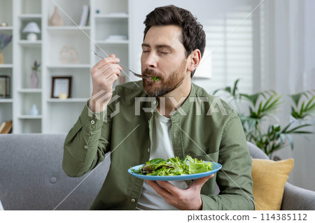 A bearded man eating a healthy green salad at home, seated comfortably in a stylishly furnished living room surrounded by plants and natural light. 114385112
