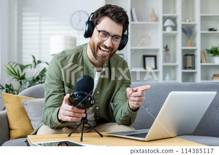 Cheerful male podcaster in headphones recording a live podcast. He is gesturing and smiling warmly at his comfortable home setup. Cheerful male podcaster in headphones recording a live podcast. He is gesturing and smiling warmly at his comfortable home setup. 114385117