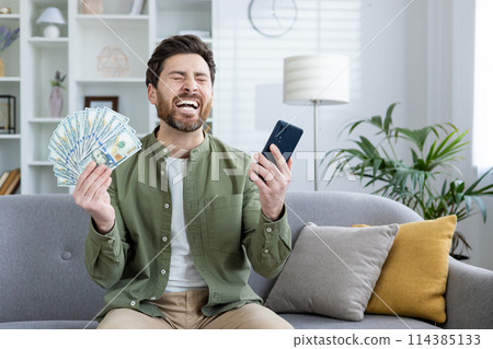 Exuberant male in a casual outfit joyously celebrating with a stack of cash and a smartphone in a cozy living room setting. Exuberant male in a casual outfit joyously celebrating with a stack of cash and a smartphone in a cozy living room setting. 114385133
