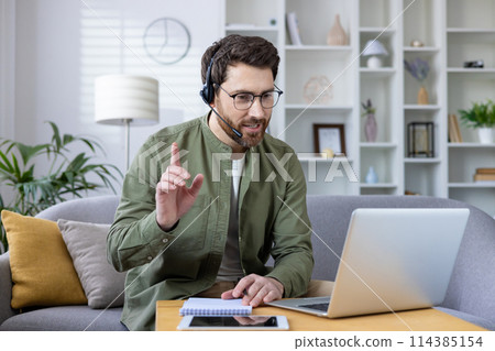 Middle-aged man with beard engaging in a virtual meeting using a headset and laptop from his well-organized home office. Middle-aged man with beard engaging in a virtual meeting using a headset and laptop from his well-organized home office. 114385154