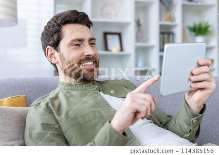 A cheerful, bearded man comfortably using a digital tablet while sitting on a couch. His relaxed posture and smile suggest he is enjoying his activity. 114385156