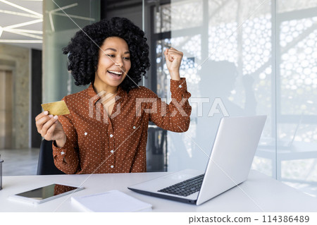 A joyful African American businesswoman celebrates a successful transaction or achievement at her modern office desk, holding a credit card and showing a victory gesture. 114386489