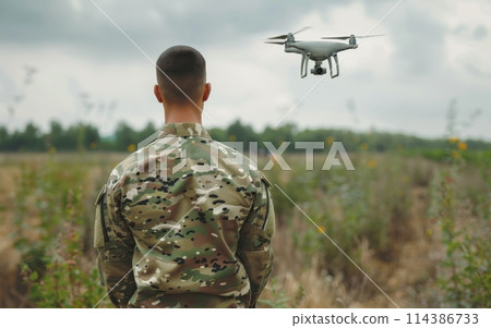 A military personnel stands focused as he controls a drone over a wildflower-dotted field, signaling a blend of natural beauty and technological advancement. Drone operator at war. 114386733
