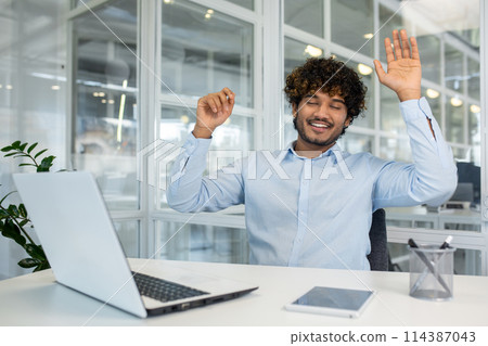 Curly islamic man in blue shirt raising arms for stretching back while sitting at workspace with closed eyes. Relaxed employee using free time for exercising and resting from sedentary work. 114387043