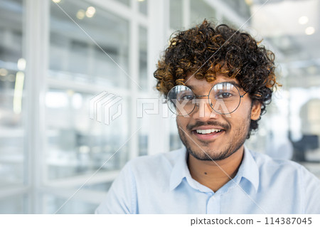 A portrait of a cheerful young man wearing eyeglasses, smiling at the camera in a bright, contemporary office environment. The image exudes positivity and confidence. 114387045