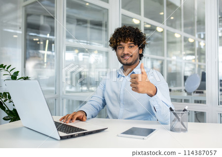 A young male professional with curly hair sits at a white desk in a bright office, giving a thumbs-up sign while working on a laptop. A young male professional with curly hair sits at a white desk in a bright office, giving a thumbs-up sign while working on a laptop. 114387057