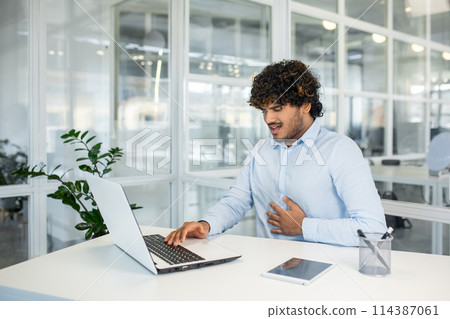 A young man in a business setting feels sudden abdominal pain while working on his laptop. The discomfort disrupts his concentration and workflow. A young man in a business setting feels sudden abdominal pain while working on his laptop. The discomfort disrupts his concentration and workflow. 114387061