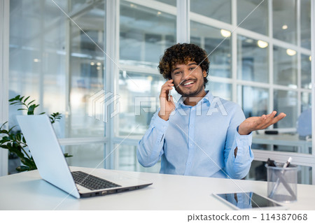 Cheerful young man with curly hair, wearing a light blue shirt, communicates on a smartphone while working at a white desk in a bright office environment. Cheerful young man with curly hair, wearing a light blue shirt, communicates on a smartphone while working at a white desk in a bright office environment. 114387068