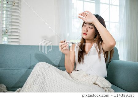 Sick woman have symptom sick high fever she checking her temperature with thermometer in living room at home, young female suffering from flu cold and fever, health care and medical concept 114389185