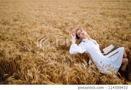 Young woman in white linen dress and hat enjoying a sunny day in a golden wheat field. Summer, beauty. 114389432