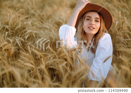 Young woman in white linen dress and hat enjoying a sunny day in a golden wheat field. Summer, beauty. 114389490