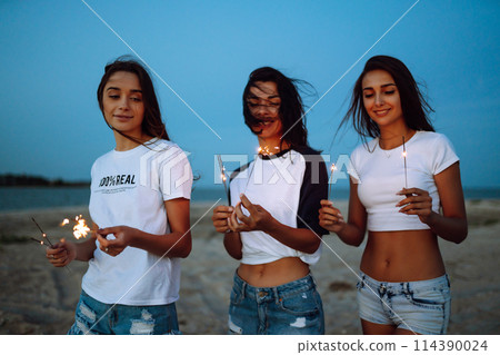 Three beautiful girls celebrating, holding sparklers on the beach at night. . Summer holidays, vacation. Three beautiful girls celebrating, holding sparklers on the beach at night. . Summer holidays, vacation. 114390024
