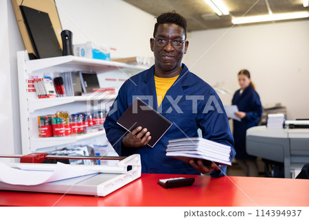 Excited middle-aged African American male typographer in uniform showing notepads in the typography Excited middle-aged African American male typographer in uniform showing notepads in the typography 114394937